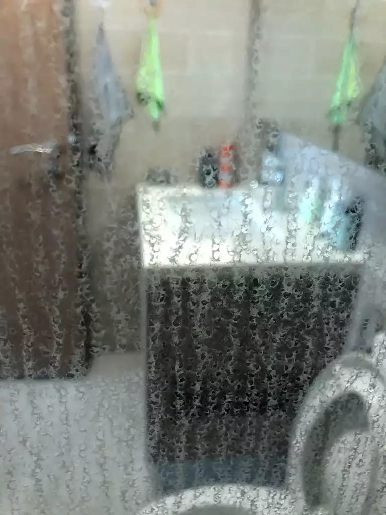 Dense spotting and vertical trails across shower glass, with bathroom fittings visible behind it